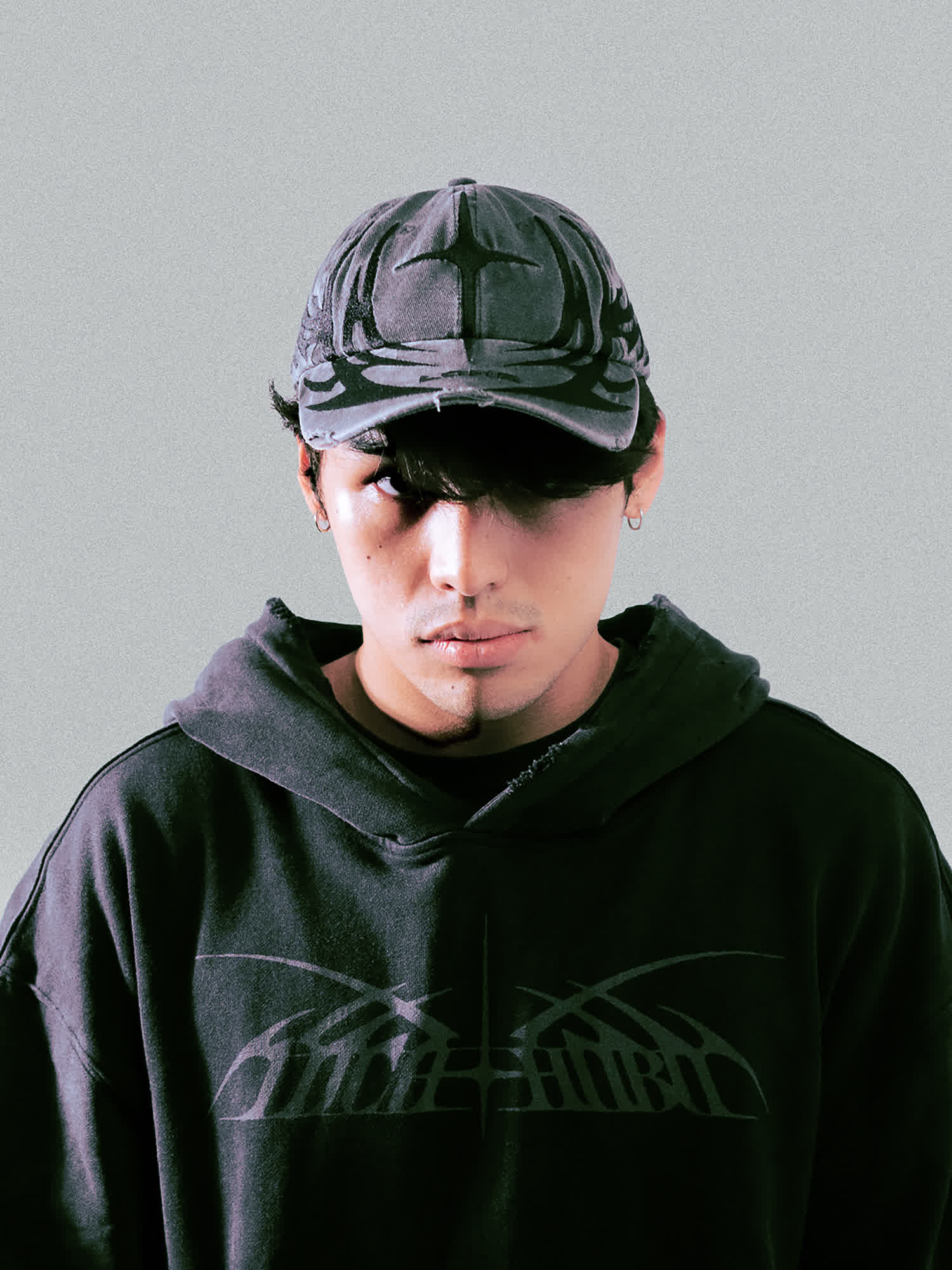 A model looks directly at the camera, wearing the PIERCE Cap — a distressed, washed-grey baseball cap featuring a large, tonal cross-like graphic on the front.