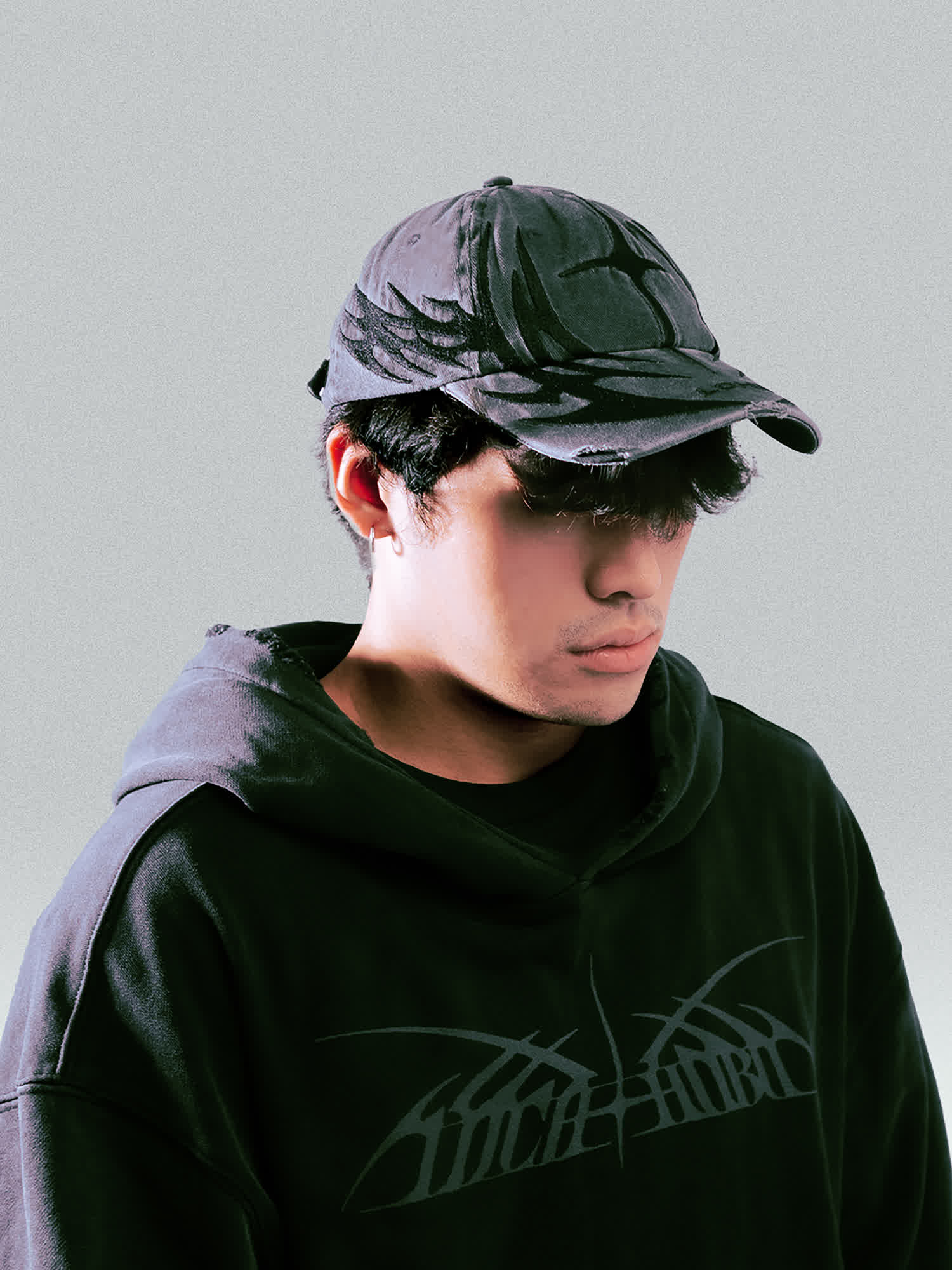 A side view of a model looking down while wearing the PIERCE Cap, which features a distressed, washed-black finish and a large, tonal graphic wrapping around the side.